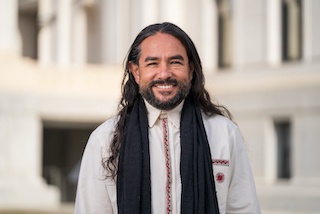 Kilo wearing a white shirt and black scarf. His is smiling at the camera